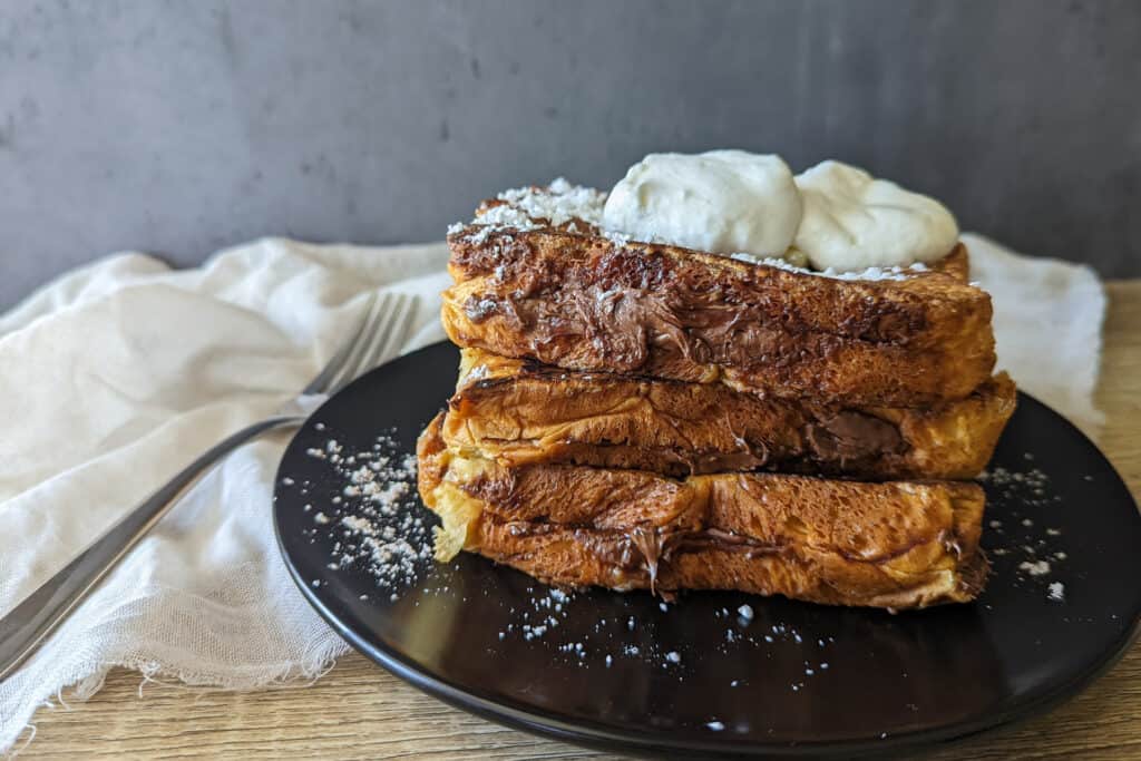 A stack of Nutella stuffed brioche french toast topped with butter, powdered sugar, and whipped cream