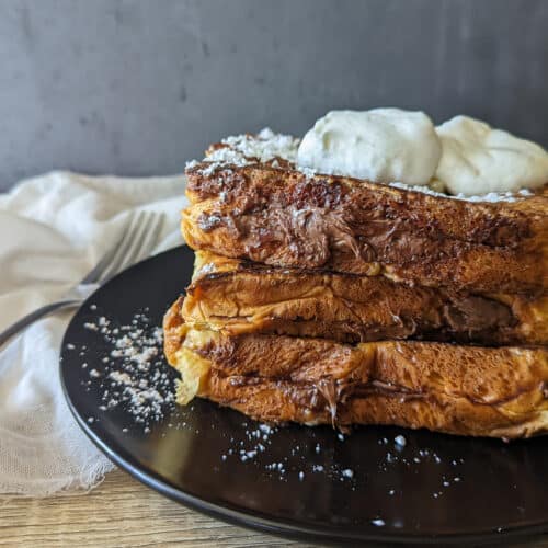 A stack of Nutella stuffed brioche french toast topped with butter, powdered sugar, and whipped cream