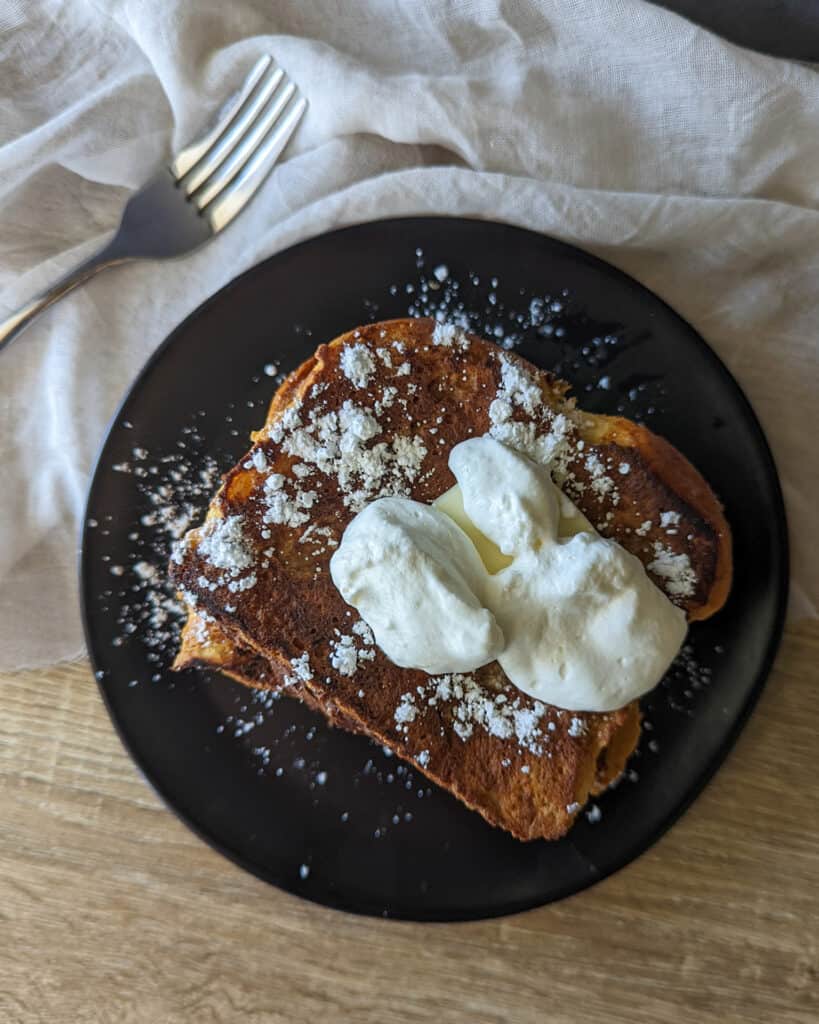 Brioche french toast topped with butter, powdered sugar, and whipped cream on a plate