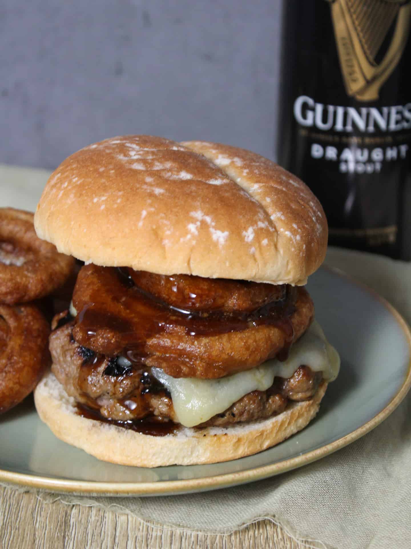 A freshly made Guinness burger topped with white cheddar, crispy onion rings, and Guinness glaze with a can of Guinness beer.