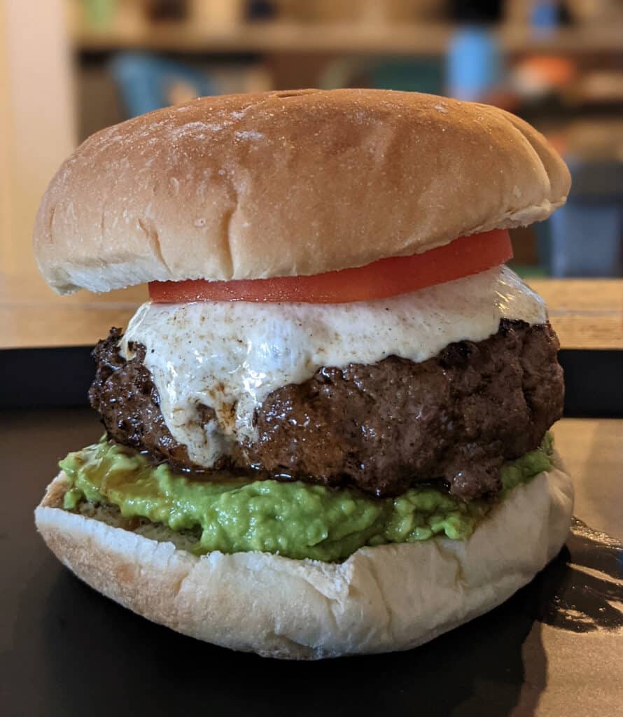 A juicy burger topped with melty mozzarella cheese, fresh tomato, and mashed avocado on a soft bun.