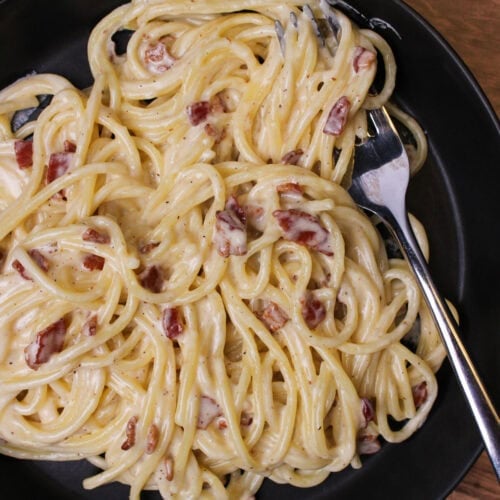 A black bowl of spaghetti carbonara with bacon and a fork.