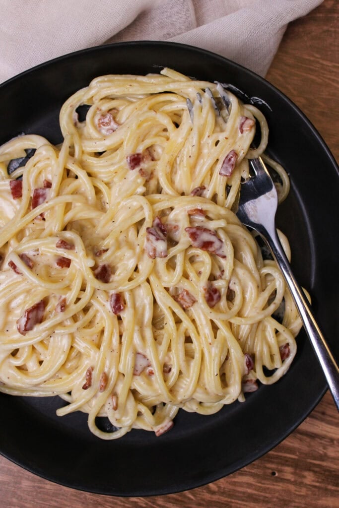 A black bowl of spaghetti carbonara with bacon and a fork.
