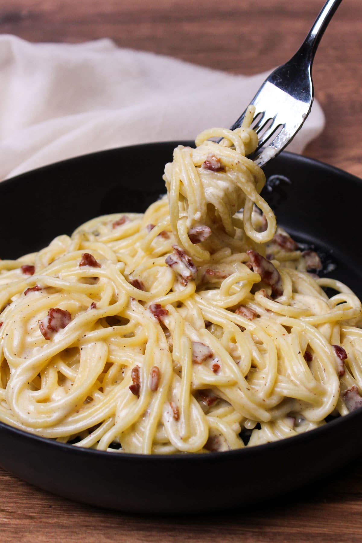 Creamy spaghetti carbonara in a black stone bowl being picked up with a fork.