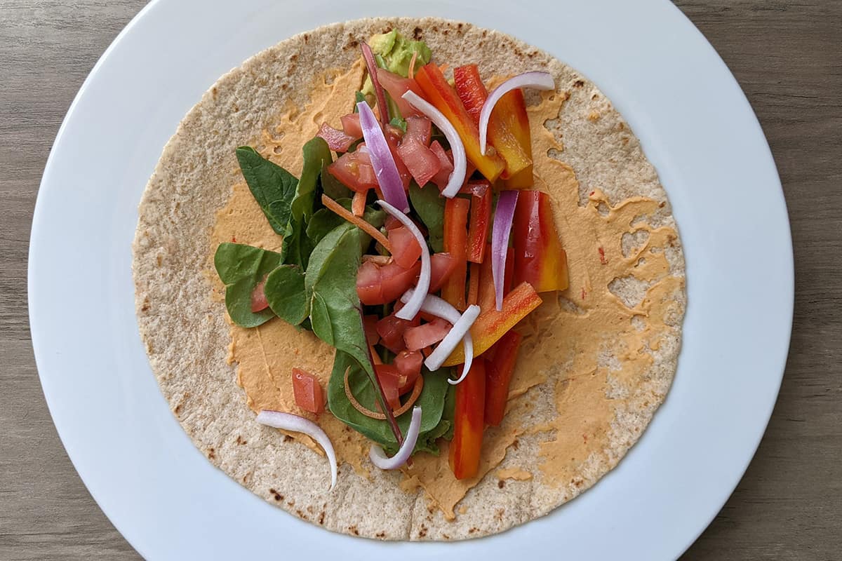 A whole wheat veggie wrap with hummus spread, avocado, lettuce, tomato, red onion, bell pepper, and carrots.