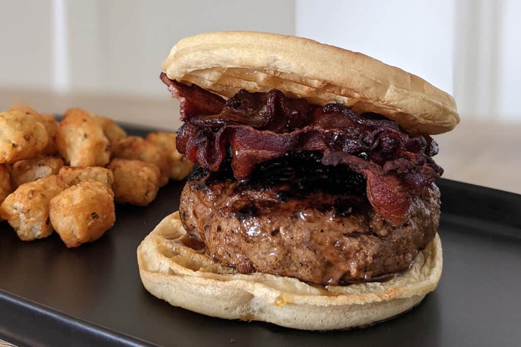 A burger with maple bourbon bacon, maple bourbon glaze, and honey butter served between two waffles with a side of tater tots.