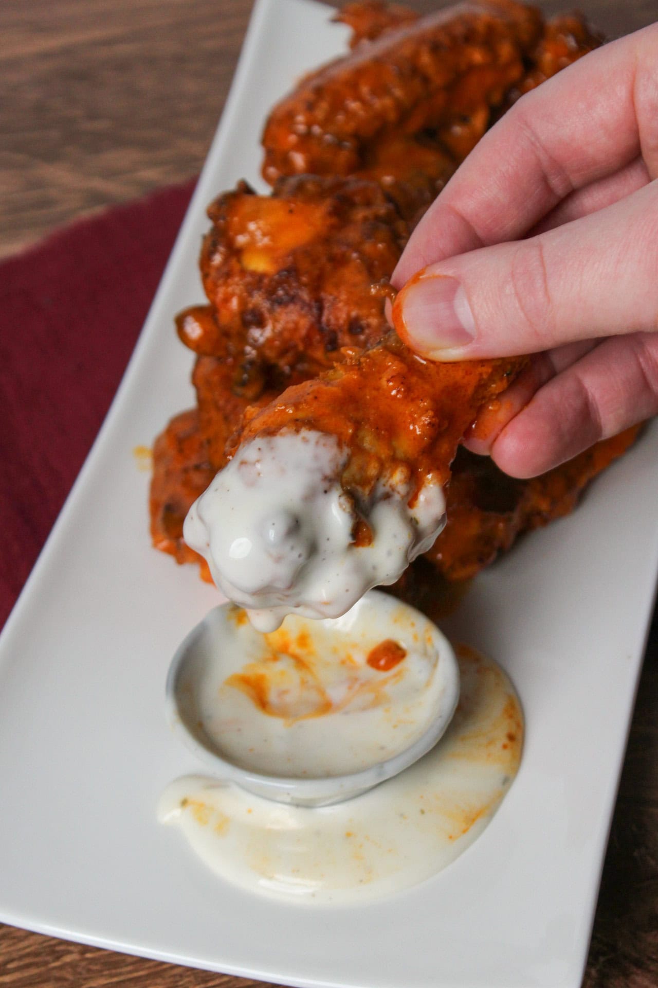 A saucy, messing buffalo chicken wing being dipped into a small bowl of ranch with the ranch overflowing.