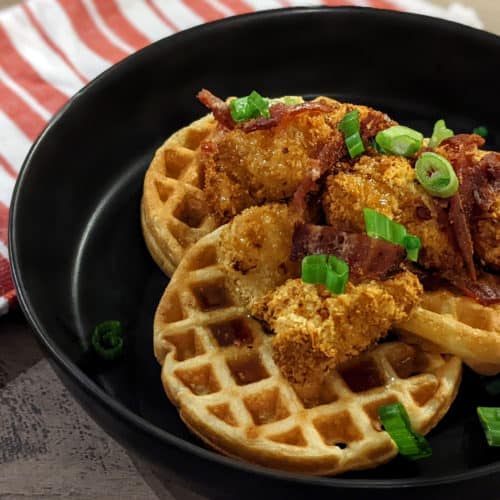 A plate with mini waffles and fried chicken nuggets, topped with bacon bits, scallions, and maple bourbon syrup.