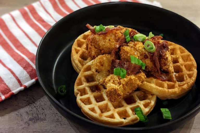 A plate with mini waffles and fried chicken nuggets, topped with bacon bits, scallions, and maple bourbon syrup.