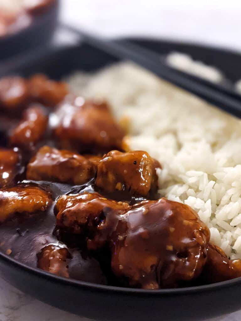 A closeup of crispy chicken in a sticky general tso's sauce served with white rice on the side