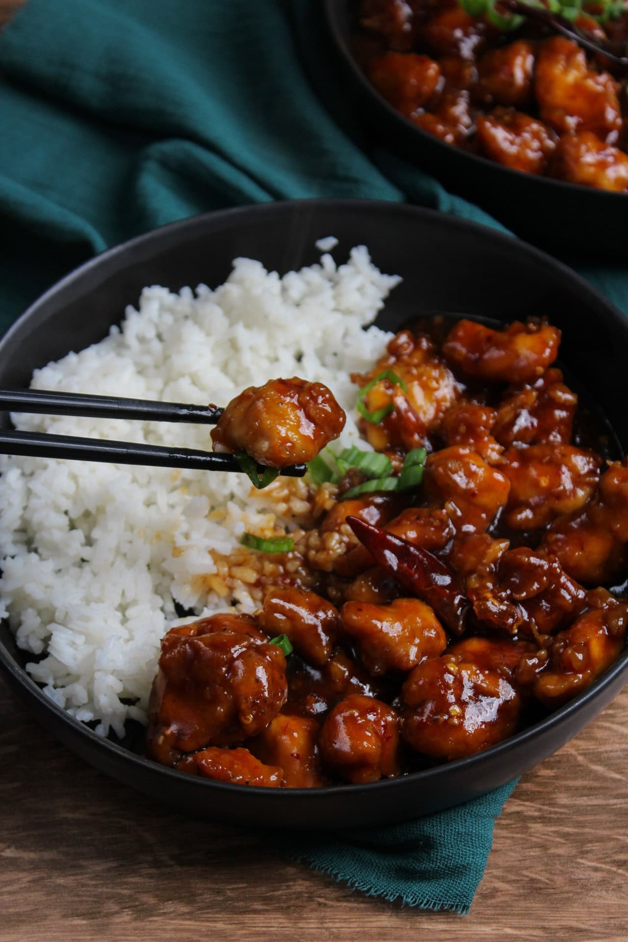 Saucy, spicy, general tso's chicken served with rice. A piece is being picked up with chopsticks. 