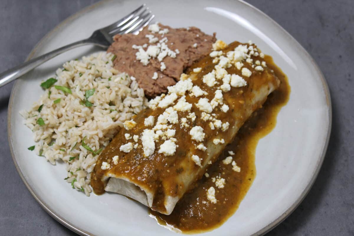 A wet burrito with green sauce and carnitas on a plate with rice and beans