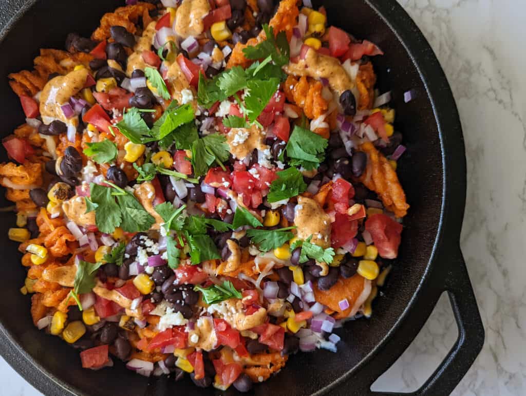 A cast iron skillet full of sweet potato waffle fries topped with cheese, beans, and produce