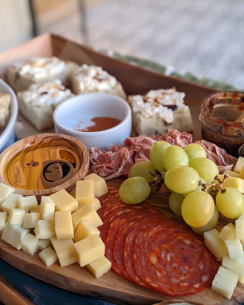 A charcuterie board with bread, dipping oils, cheddar, manchego, soppressata, prosciutto, green grapes, jam, and spices
