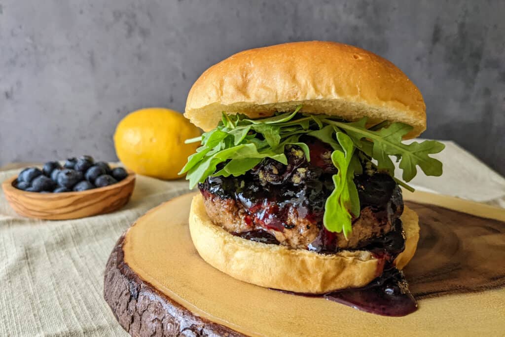 A burger on a brioche bun topped with blueberry preserves, lemon zest, goat cheese, and arugula