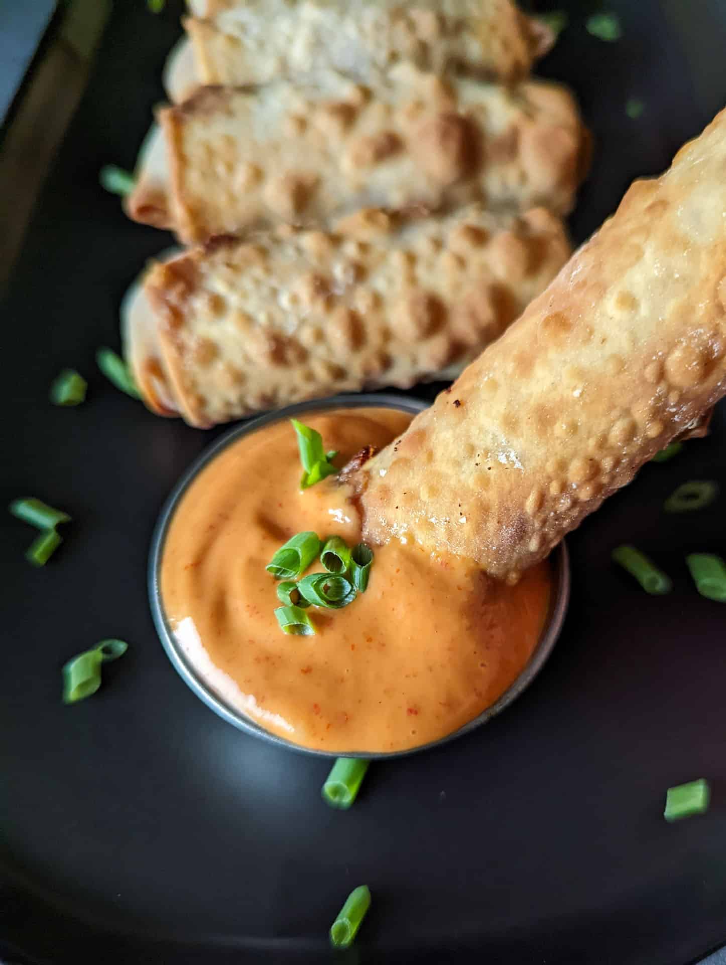 A crispy cheeseburger egg roll being dipped into spicy sriracha mayo.