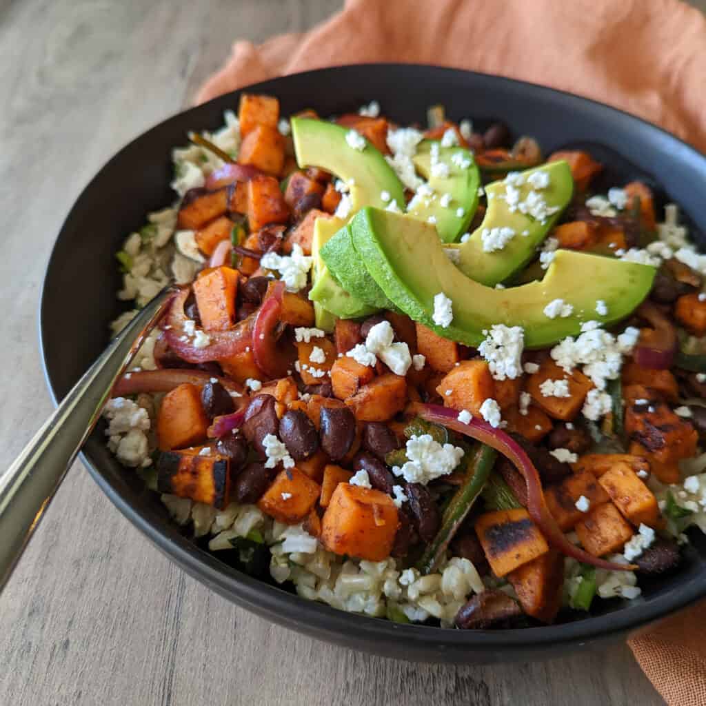 A black bean sweet potato rice bowl topped with fresh avocado slices and crumbled goat cheese