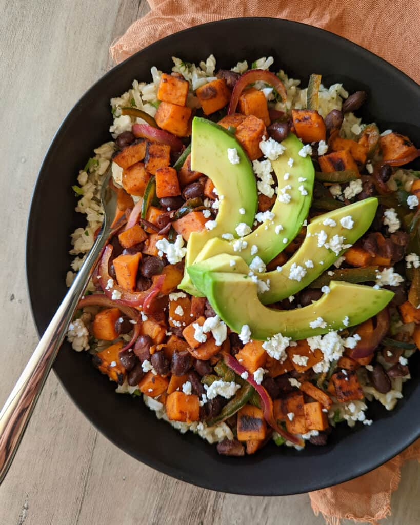A lunch bowl of sweet potatoes, black beans, brown rice, veggies, and fresh avocado topped with goat cheese