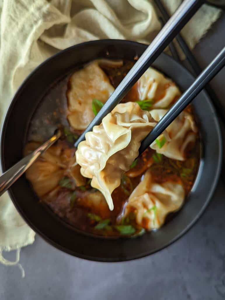 A homemade pork dumpling being lifted out of a bowl of spizy szechuan dumpling soup