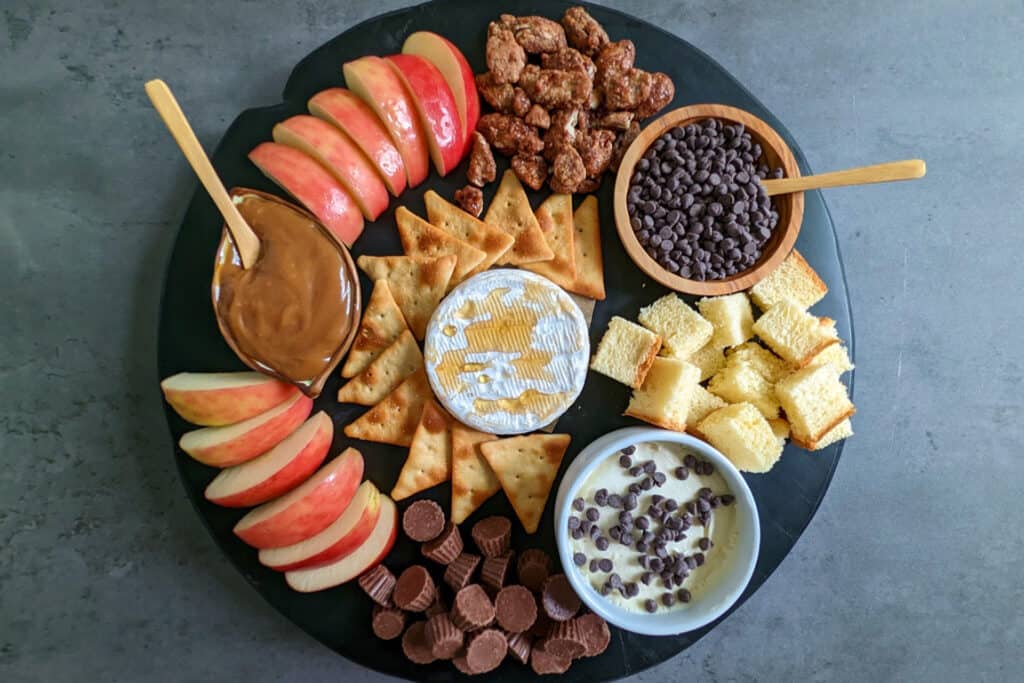 A sweet charcuterie board with apples, caramel, crackers, brie, mascarpone, brioche, candied nuts, and chocolates
