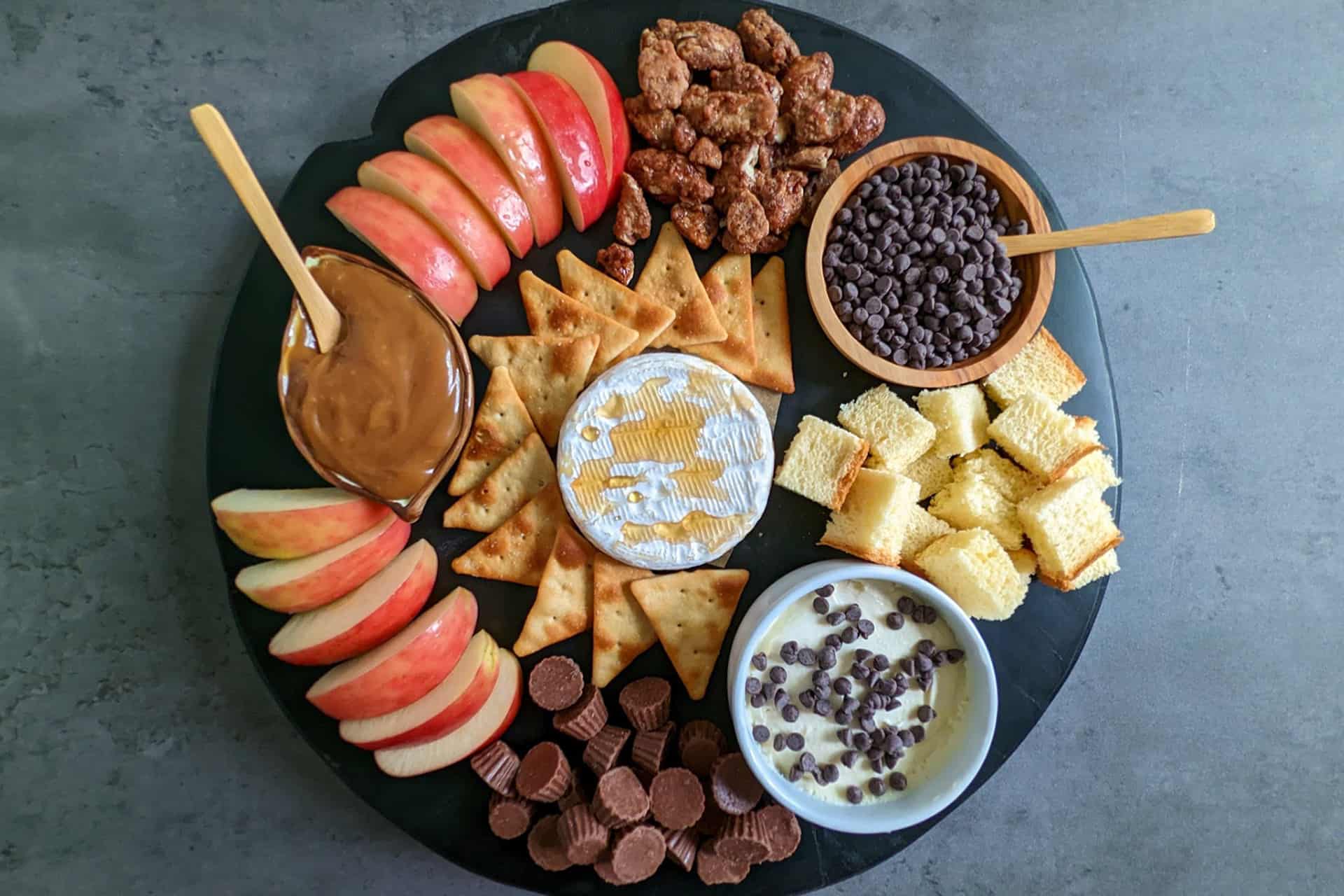 A sweet charcuterie board with apples, caramel, crackers, brie, mascarpone, brioche, candied nuts, and chocolates