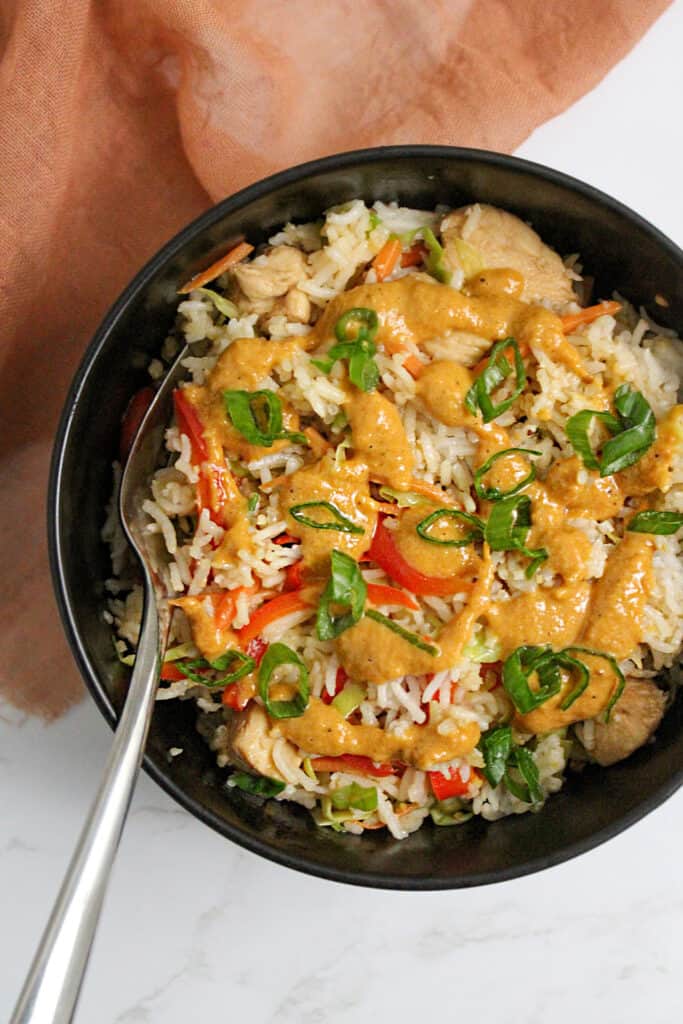 A bowl of jasmine rice with Thai chicken, veggies, and peanut sauce served with a fork