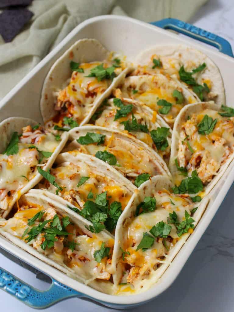 A Staub casserole dish full of baked chicken tacos with flour tortillas and cheese garnished with cilantro