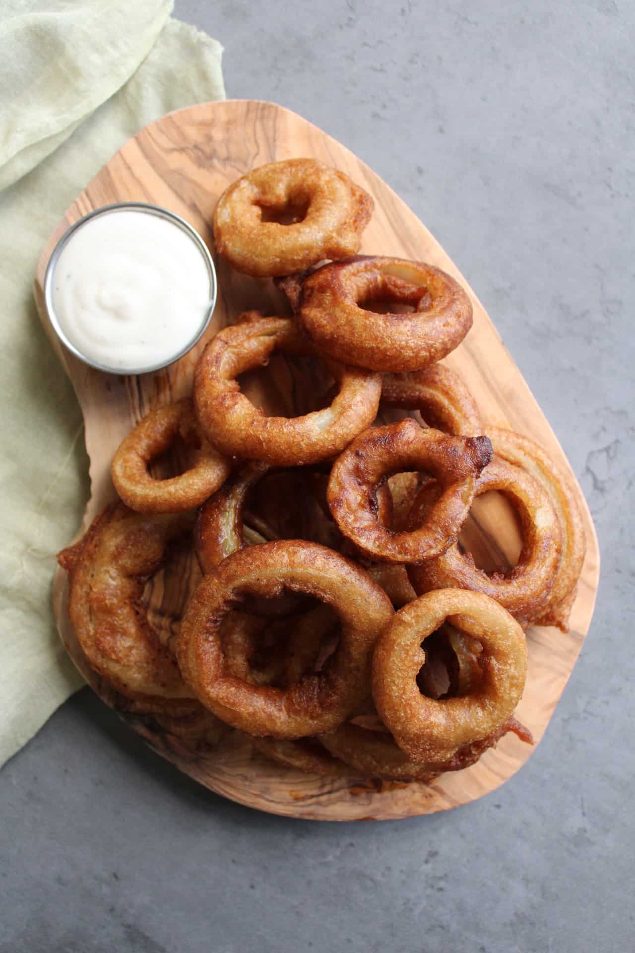 A serving board piled with homemade beer battered onion rings served with a side of ranch.
