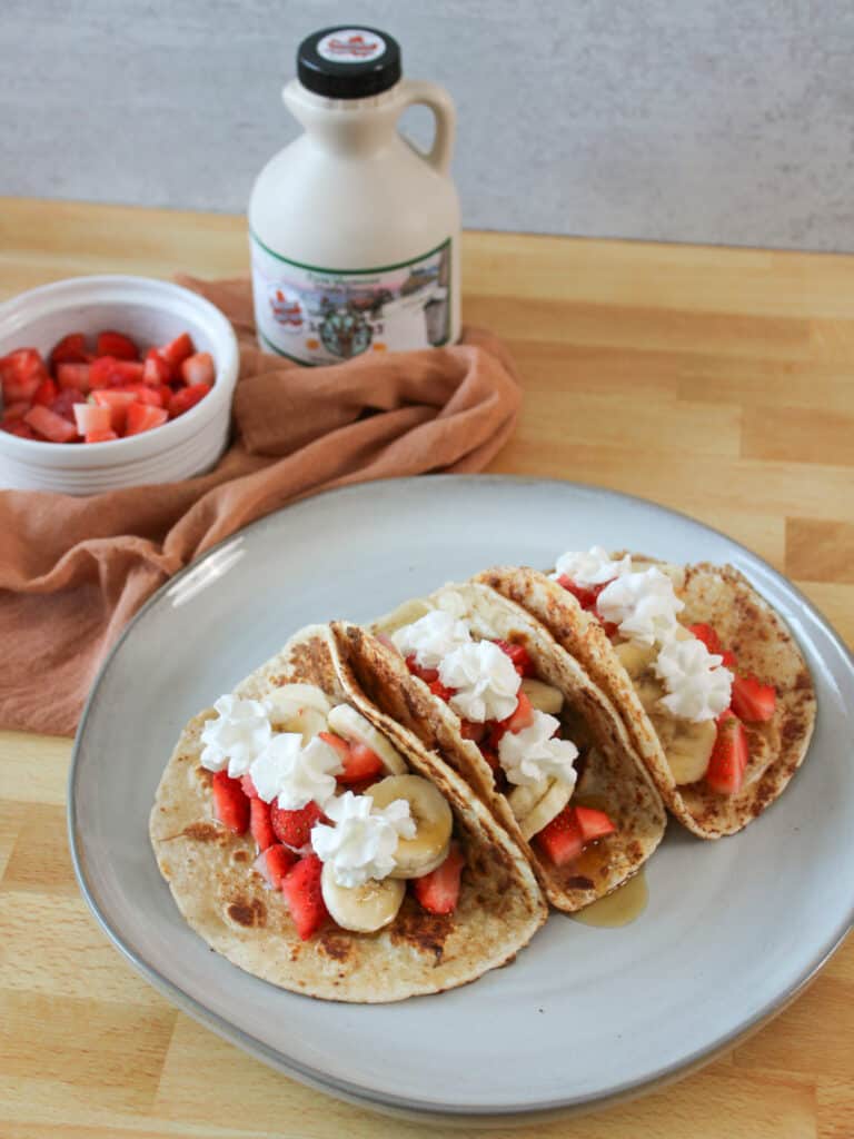 A plate of French toast breakfast tacos with maple syrup and a bowl of strawberries.