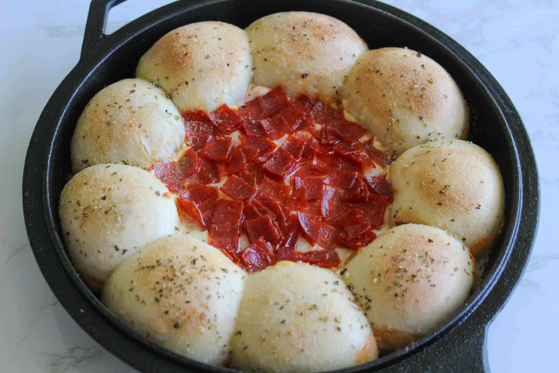 A cast iron skillet of pepperoni pizza dip surrounded by a ring of fluffy dinner rolls.