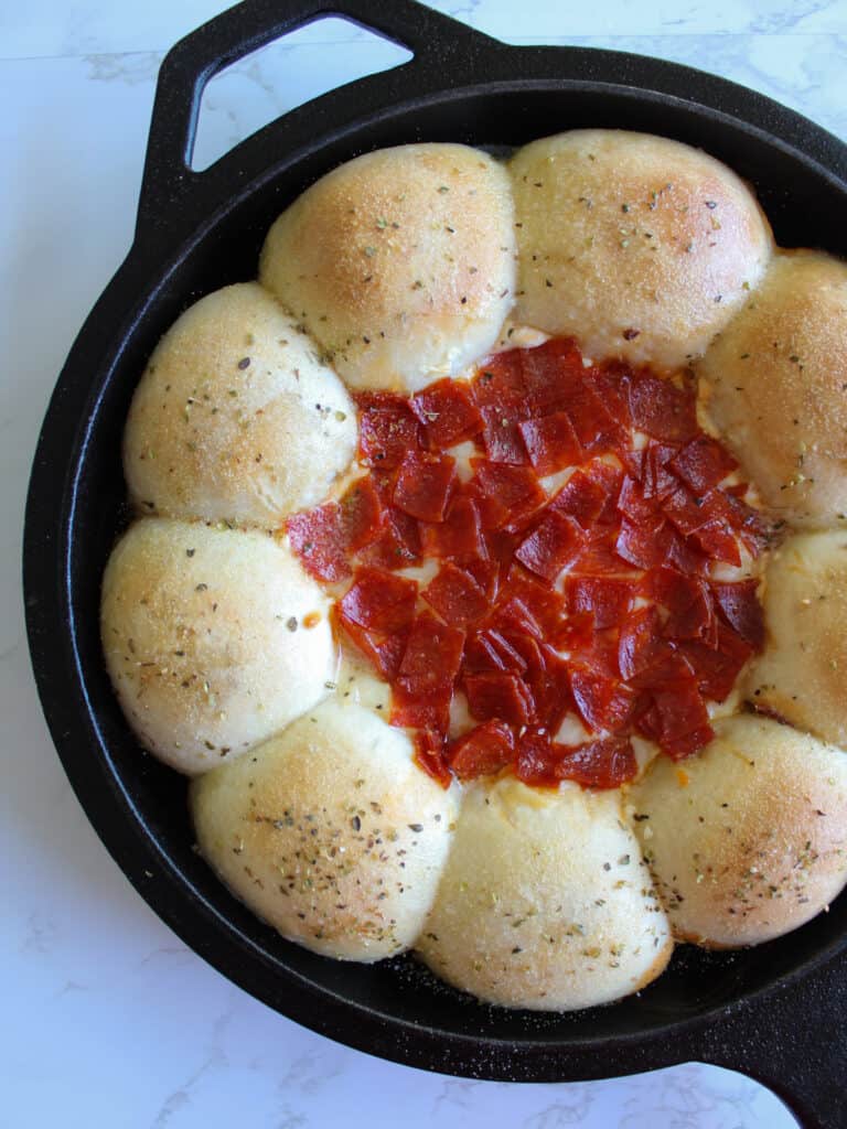 A cast iron skillet of cheesy pepperoni pizza dip with dinner rolls.