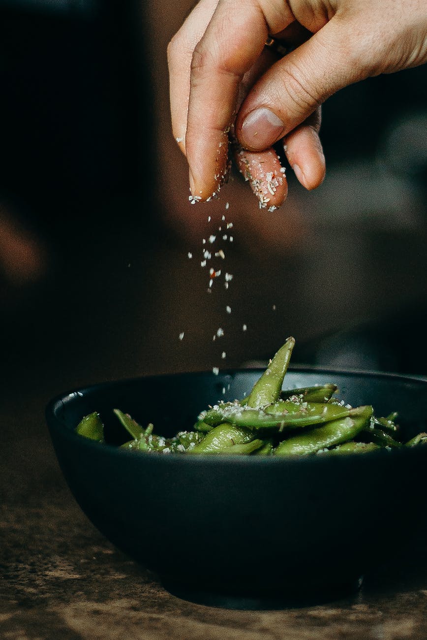 A person adding seasoning to green beans in a bowl.