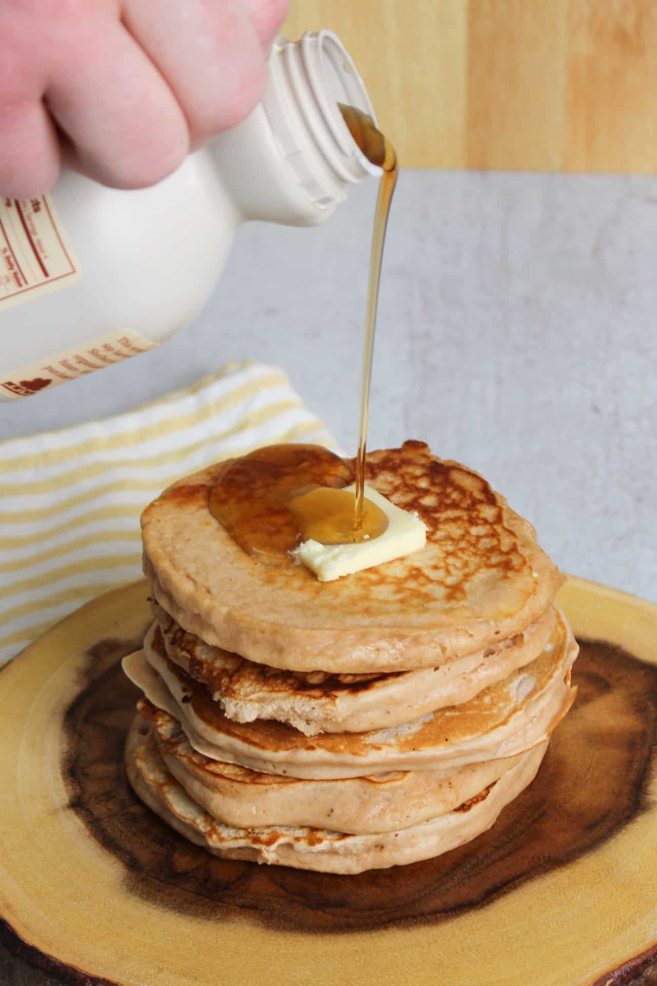 Maple syrup being poured over a stack of buttermilk protein pancakes topped with butter until it overflows down the side.