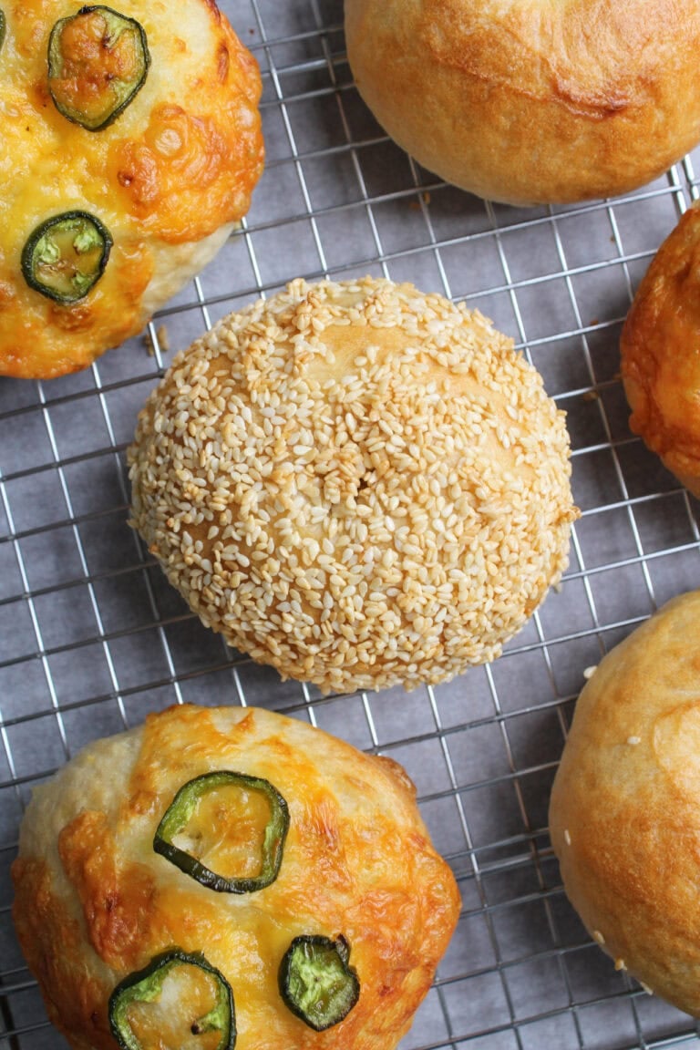 Jalapeno cheddar bagels, a sesame bagel, and plain bagels on a wire cooling rack.
