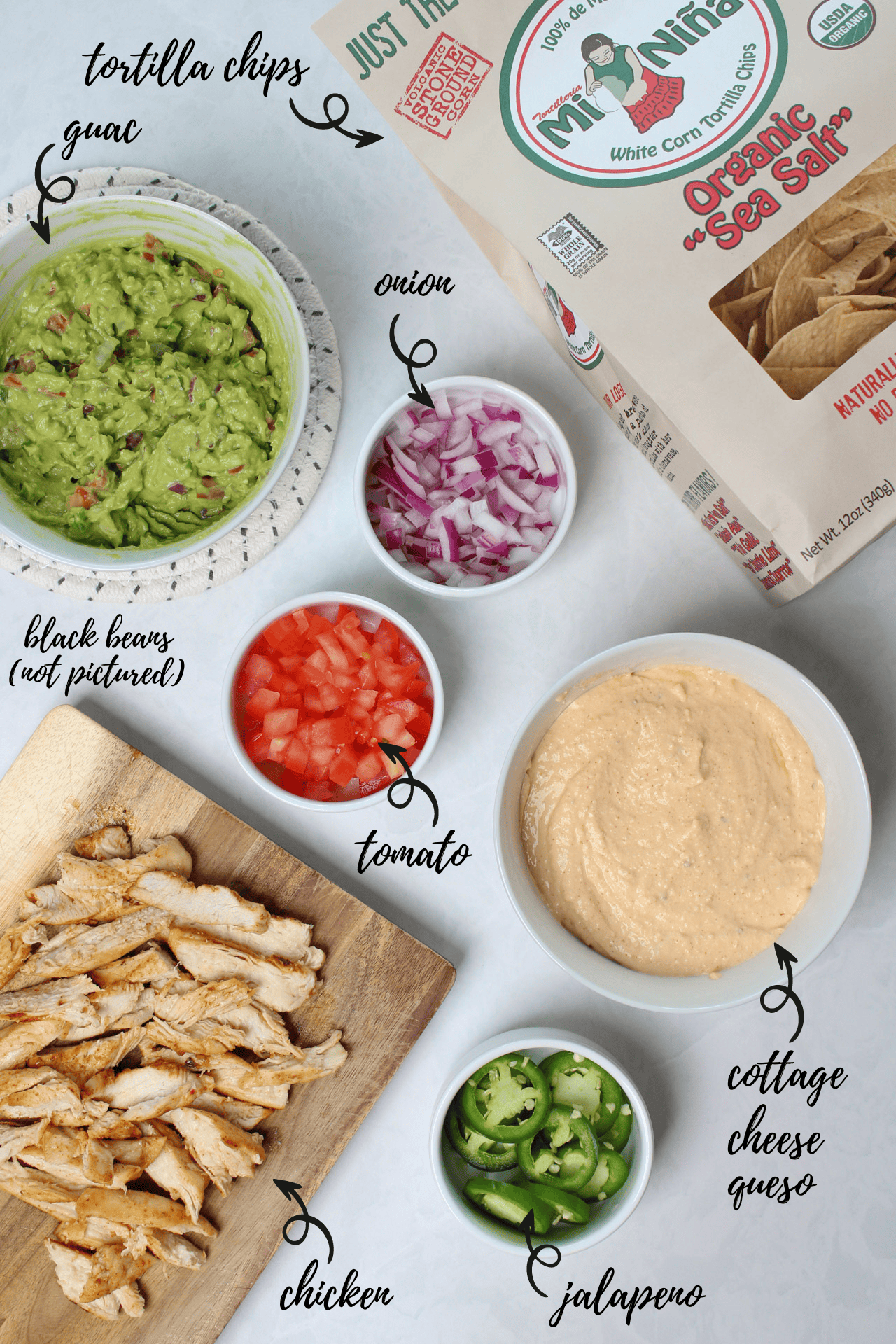 Ingredients for making high protein nachos: guac, onion, toamto, jalapeno, cottage cheese queso, chicken, black beans, and tortilla chips.