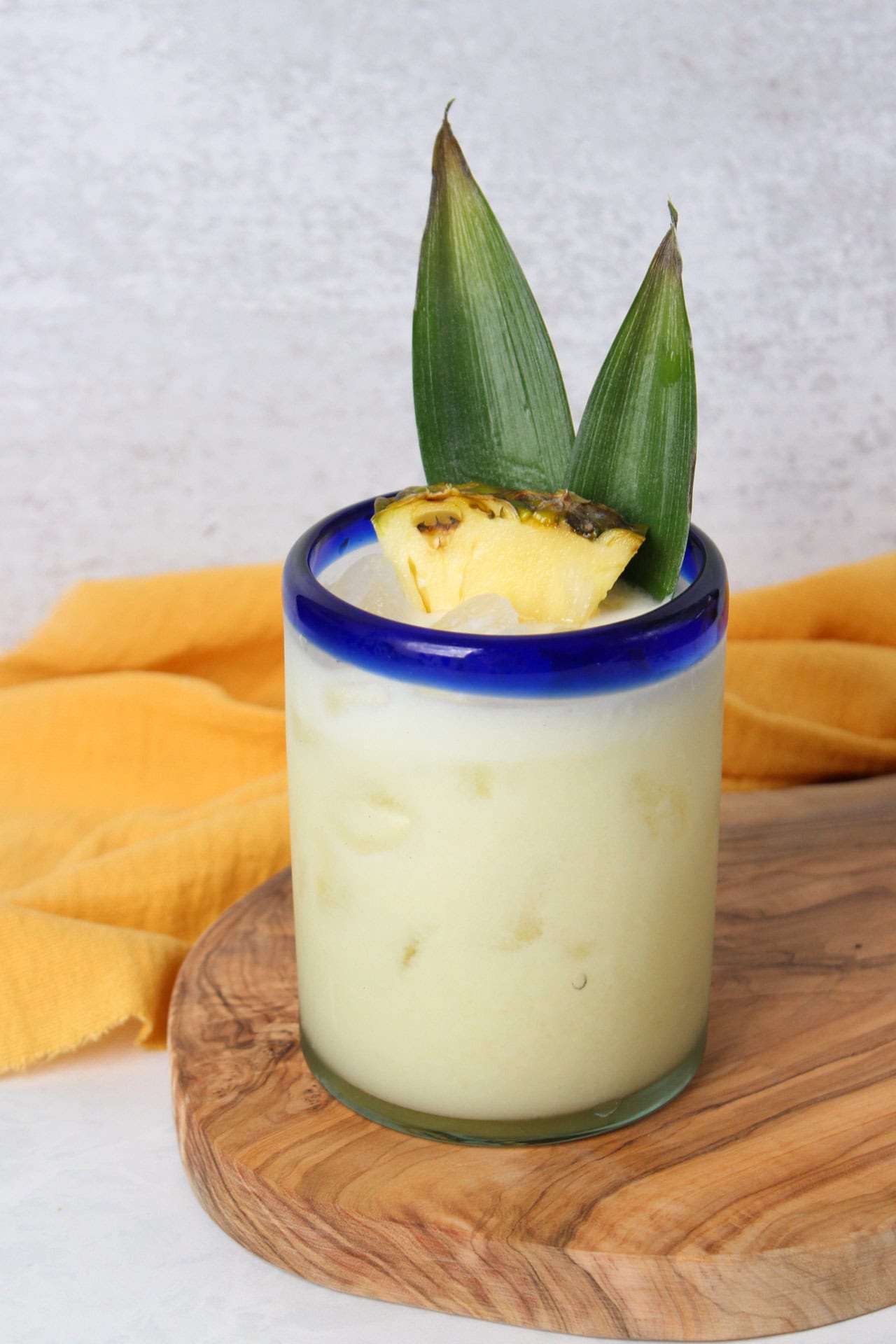 A creamy pina colada made with tequila in a clear glass with a blue rim garnished with pineapple leaves and a pineapple wedge. 
