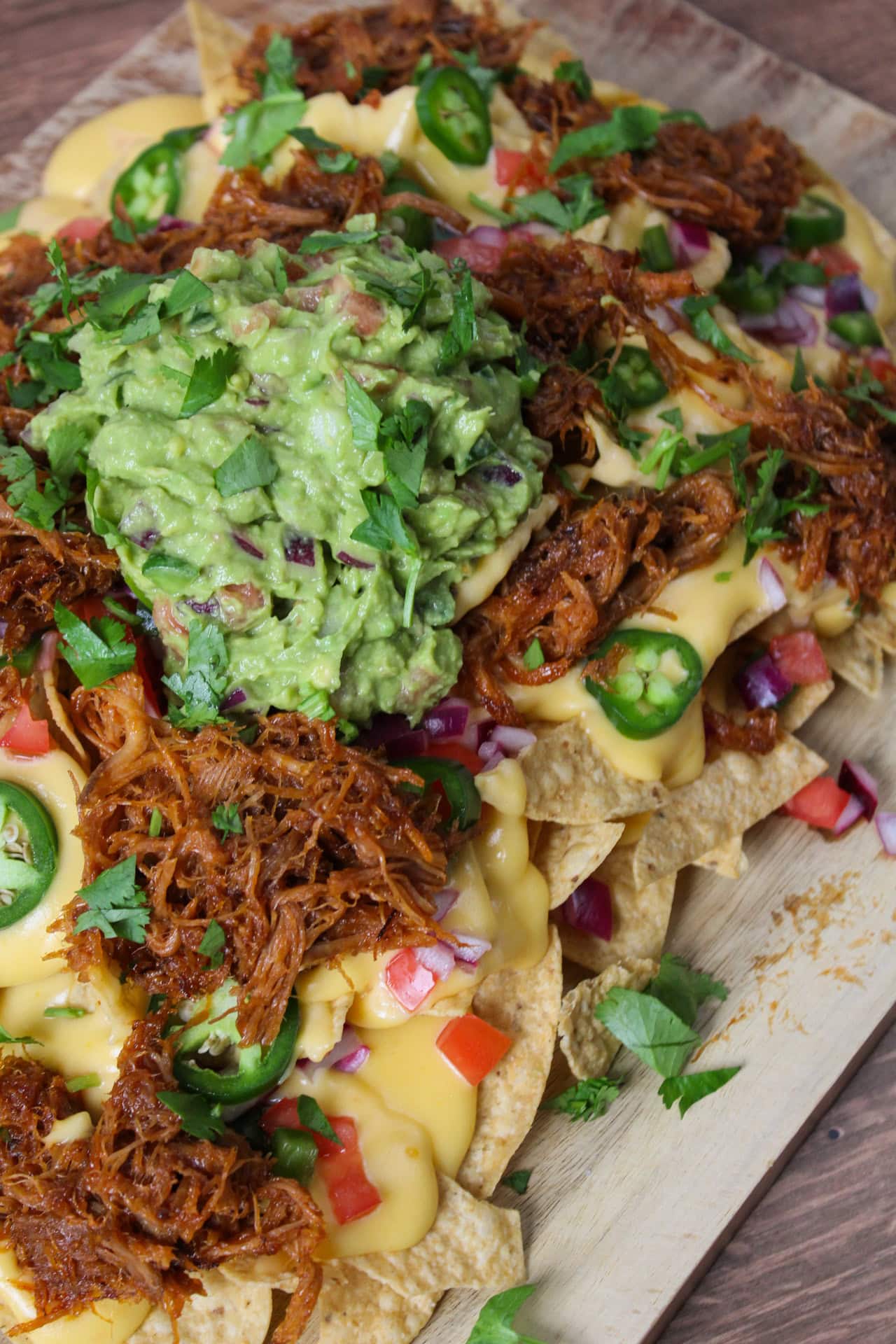 Barbecue pork nachos piled high with a cheese sauce, guaq, and other toppings on a wooden serving platter.