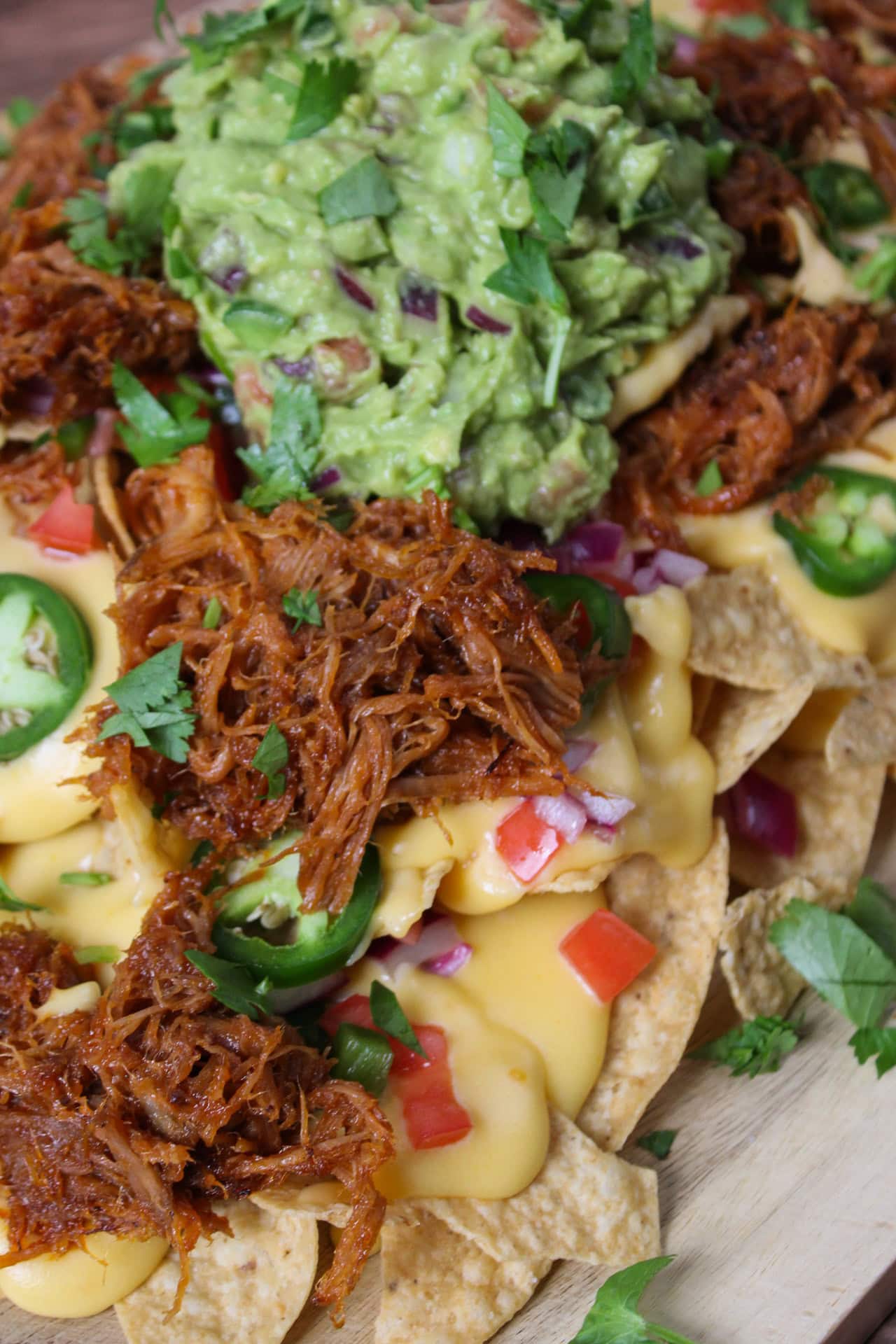 A large pile of nachos topped with pork, guacamole, cheese sauce, and pico on a wooden serving board.