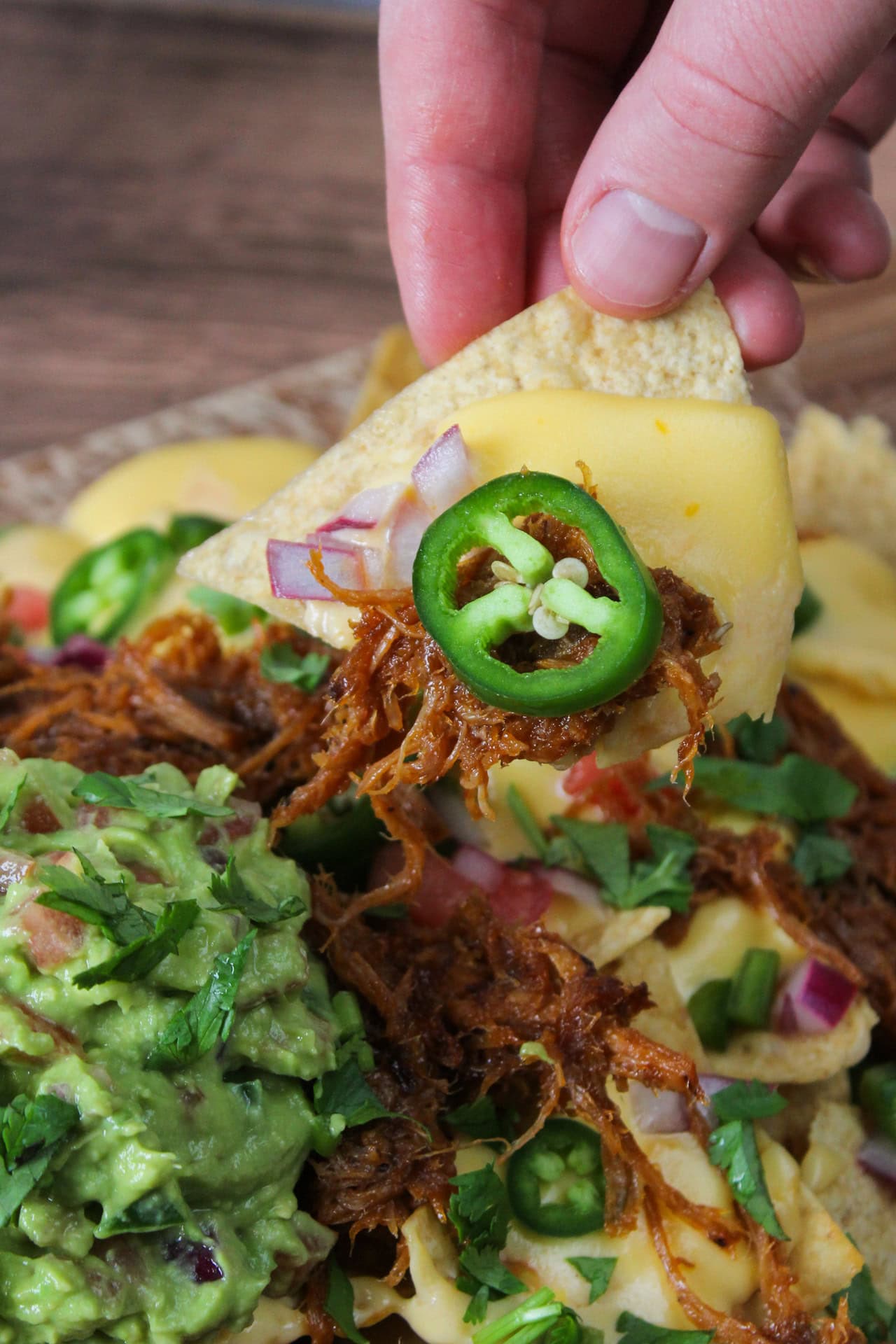 A tortilla chip with cheese sauce, pork, and jalapeno being pulled out from a pile of nachos.