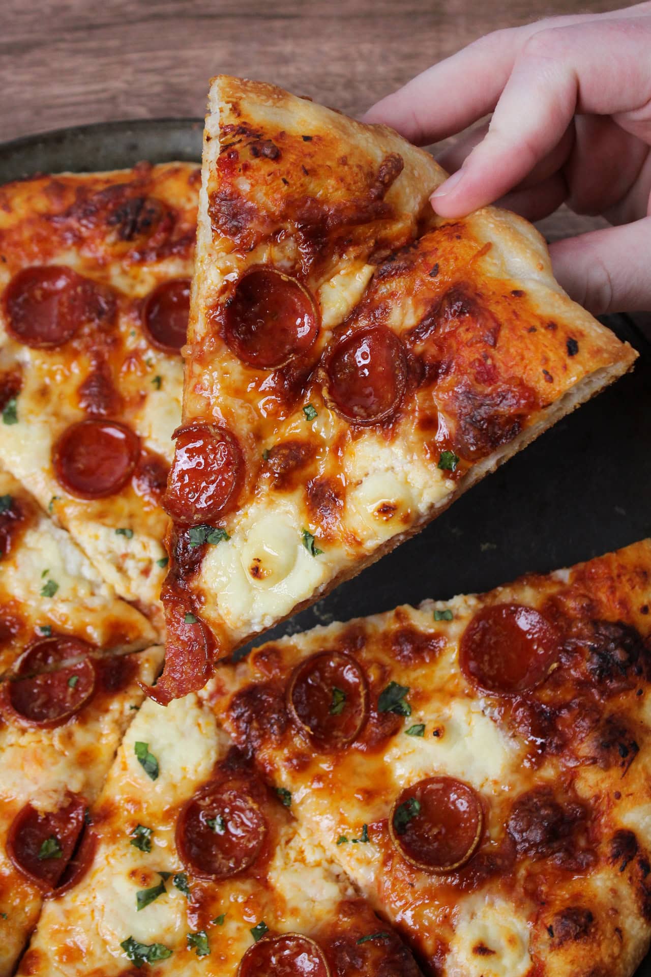 A slice of pepperoni pizza with ricotta, hot honey, and basil being held over the rest of the pizza.
