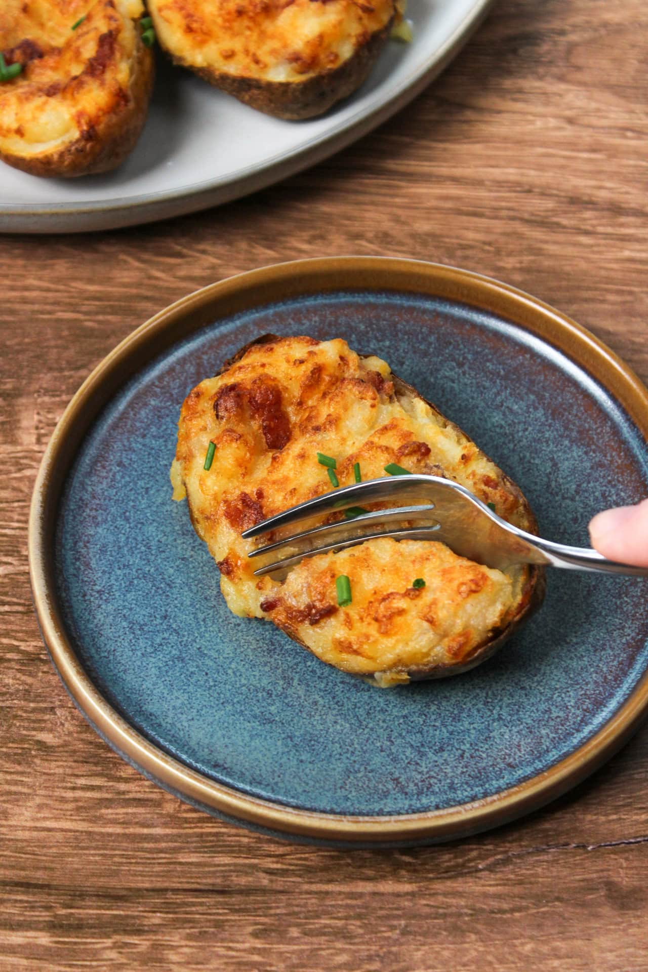 A twice baked potato filled with  bacon and cheddar cheese being cut into with a fork on a blue stoneware plate.