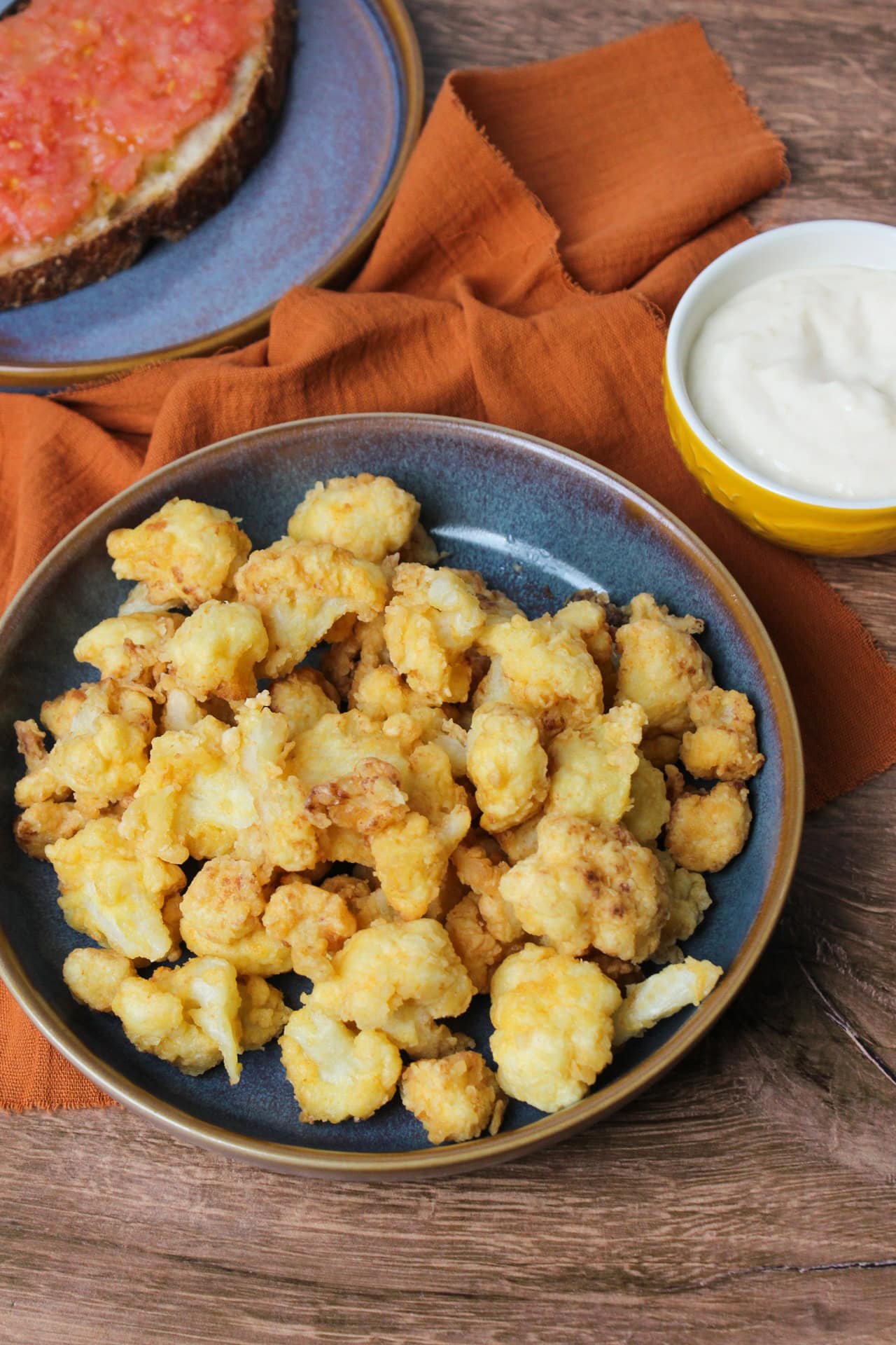 A bowl of fried cauliflower florets golden in color and served in a stoneware bowl with a side of garlic aioli.