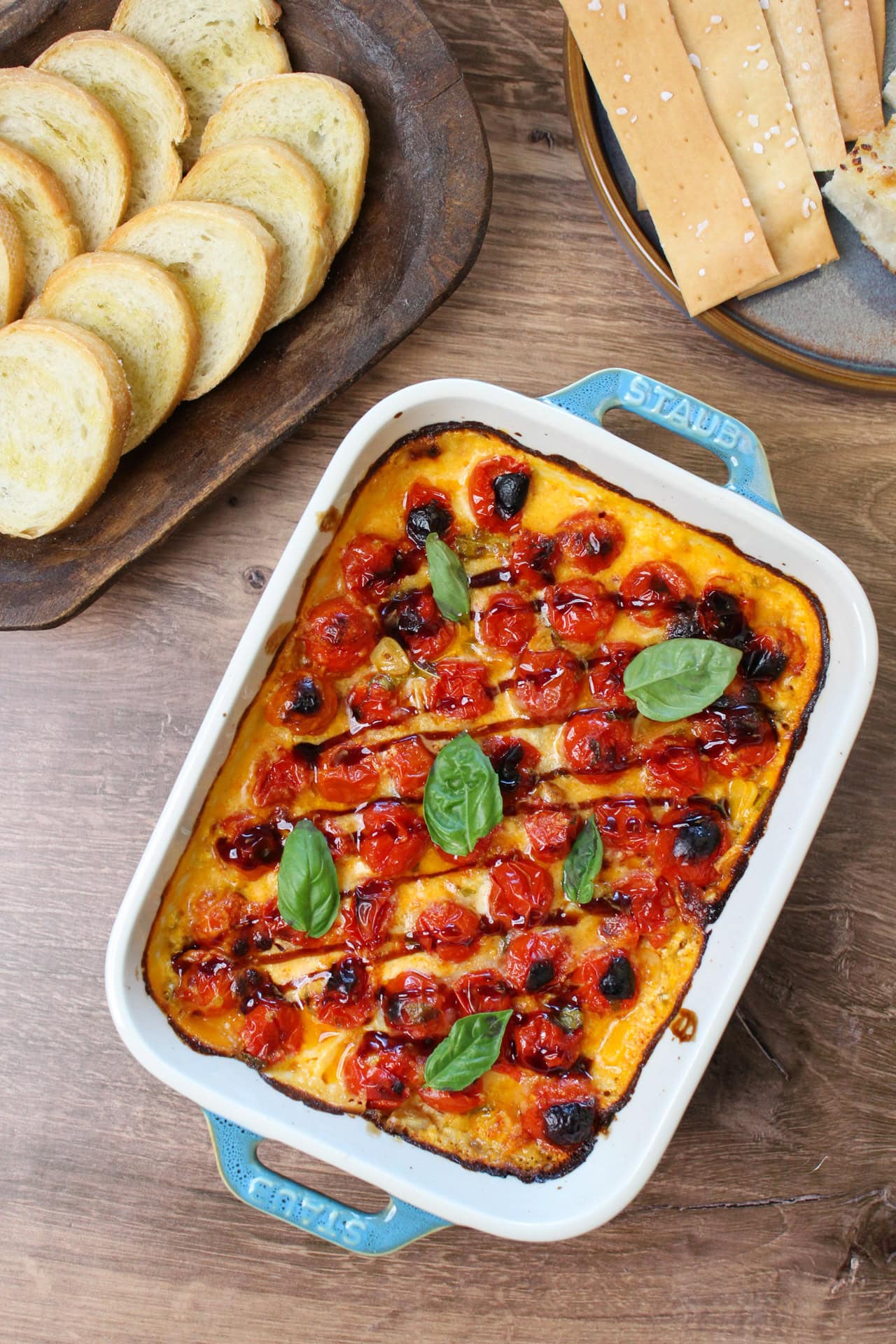 A cheesy, creamy bruschetta dip topped with fresh basil and a drizzle of balsamic glaze served with crackers and crostini.