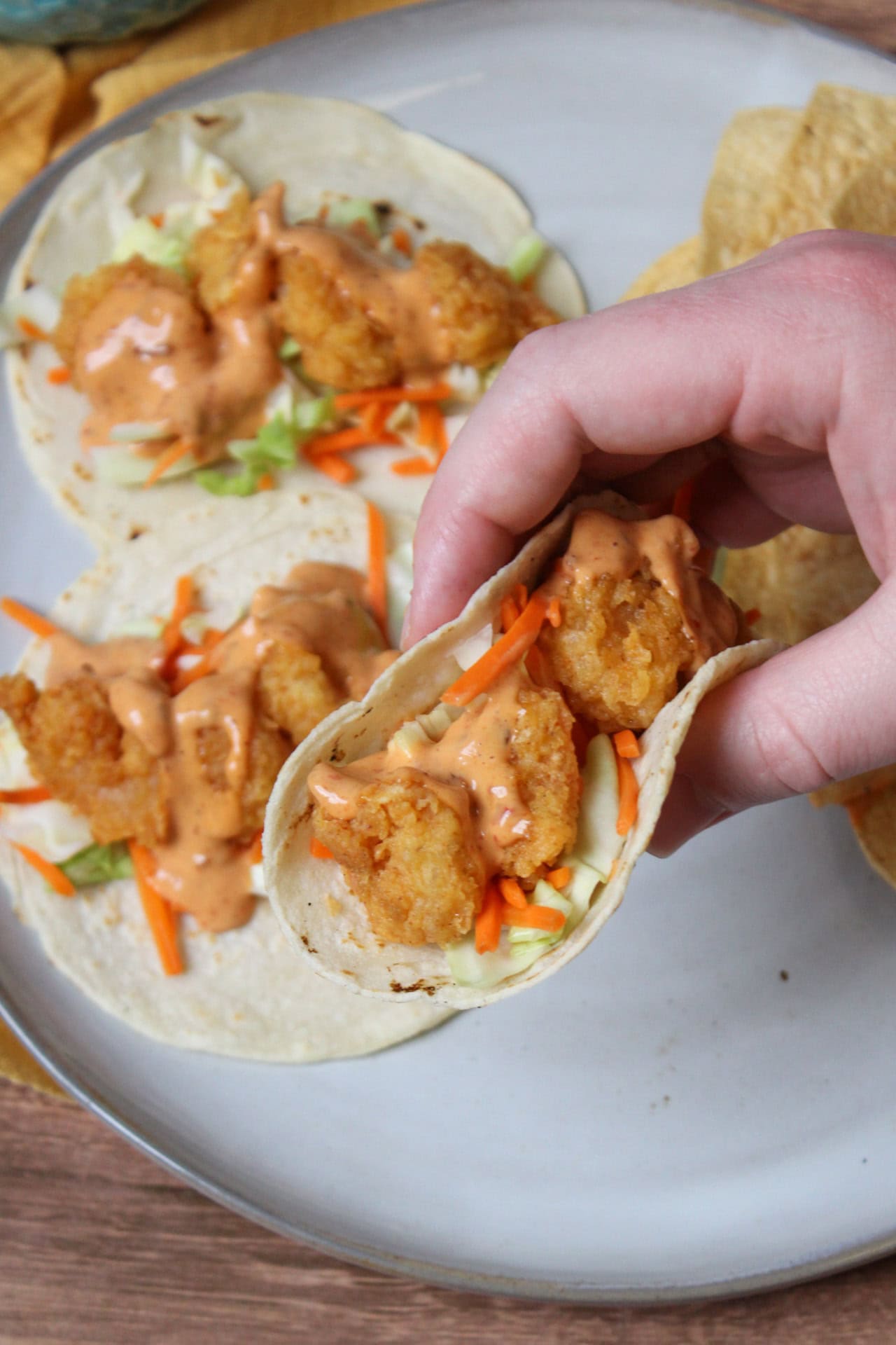 A fried shrimp taco with carrot and cabbage slaw and chipotle mayo being held above a white plate with more tacos. 