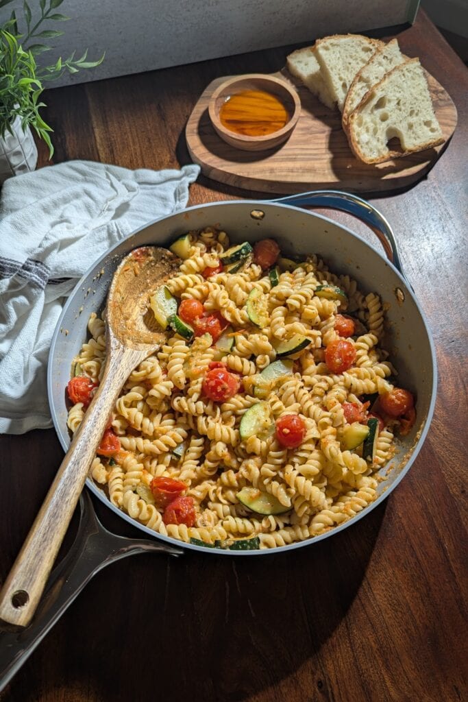 A pan of zucchini and tomato pasta with bread and dipping oils on the side.