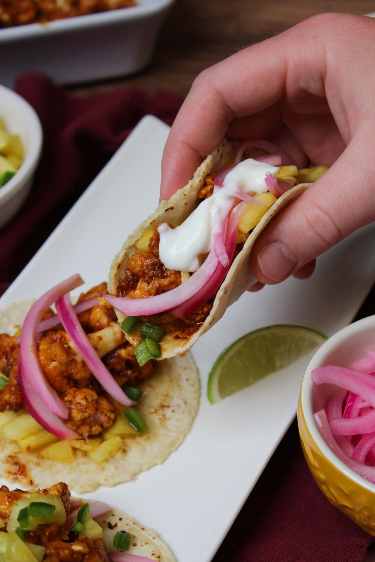 An air fryer chipotle cauliflower taco  topped with pickled red onion, a pineapple pico de gallo, and a lime crema being held over a plate of more tacos.