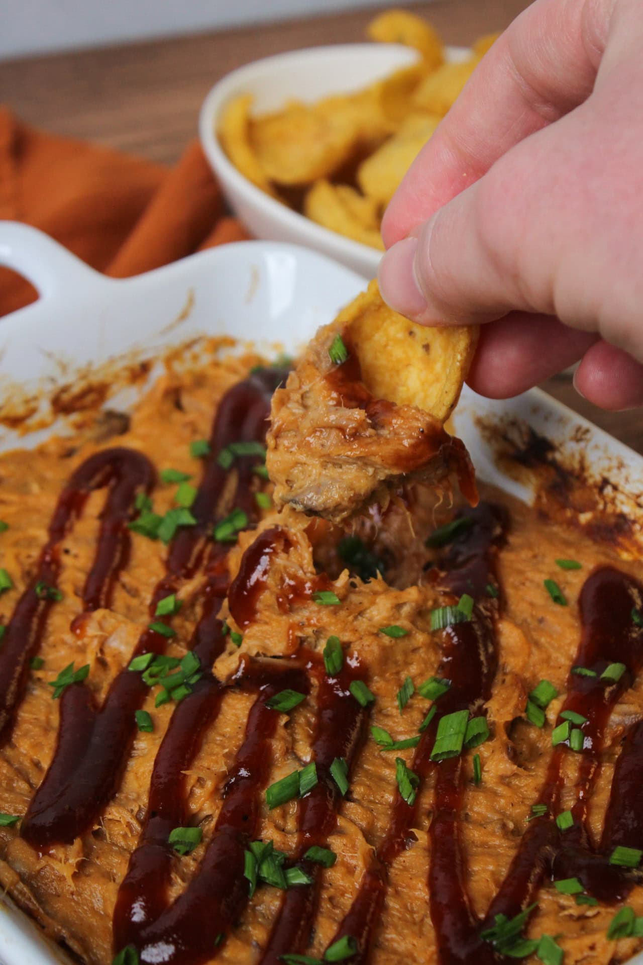 A Frito dipping into pulled pork dip that is topped with a drizzle of BBQ sauce and chives.