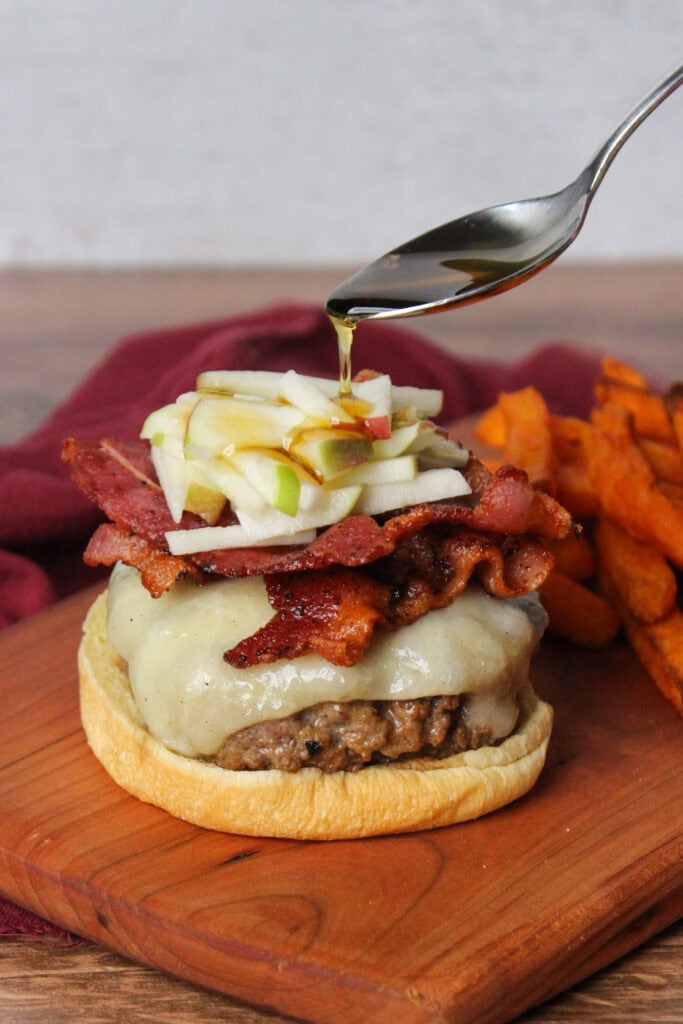 Maple syrup being poured on a burger with cheese, bacon, and apple slaw.