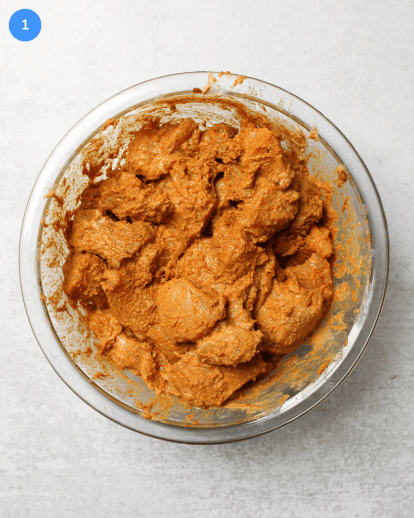 A glass mixing bowl filled with chunks of chicken marinated in yogurt, lemon juice, and spices for making chicken tikka masala.