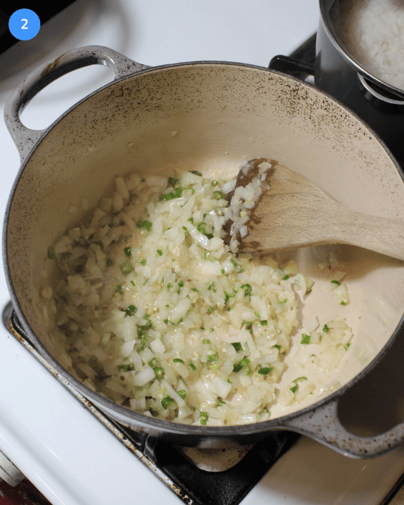 A ceramic dutch oven with onions, Thai chilies, garlic, and ginger being stirred with a wooden spatula.